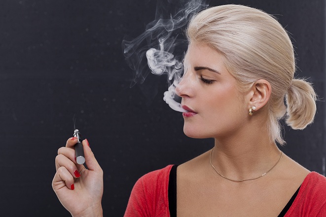 Stylish blond woman smoking an e-cigarette exhaling a cloud of smoke with her eyes closed in enjoyment, profile view on a dark background