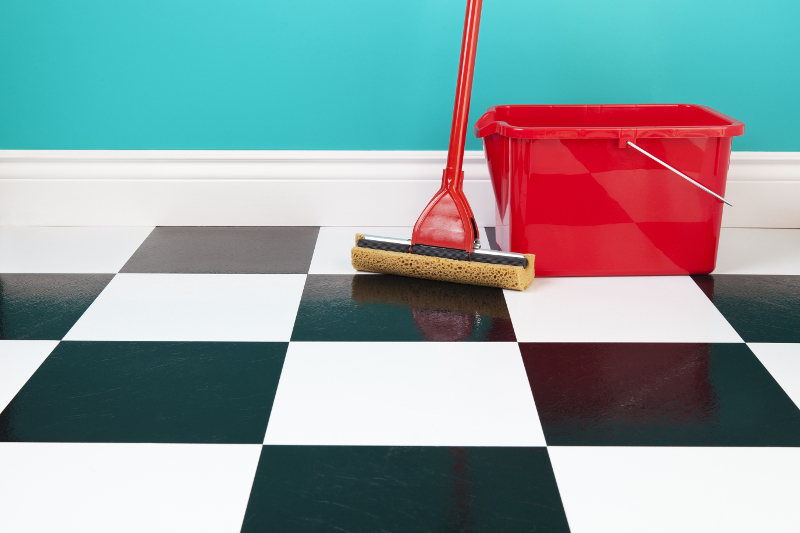 A red bucket and mop on a white and black checkered floor against a turquoise blue wall.